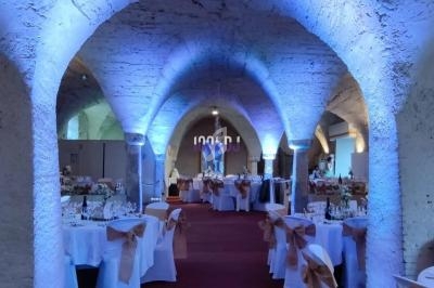 Salle voûtée éclairée en bleu, aménagée avec des tables rondes décorées pour un événement festif.