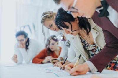 Des personnes concentrées écrivent sur des documents posés sur une table blanche dans un environnement lumineux.
