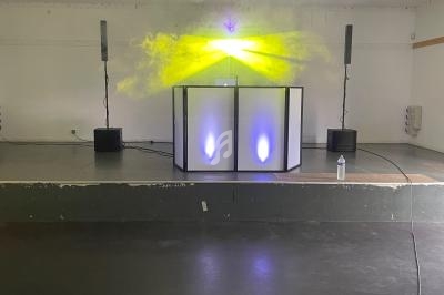 Une scène avec une installation de DJ, éclairée par des faisceaux lumineux jaunes et entourée de haut-parleurs.
