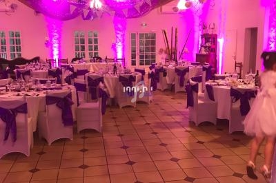 Salle de réception décorée avec des nappes blanches et des rubans violets, éclairée par une lumière rose.