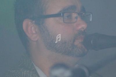 Homme barbu portant des lunettes, chantant dans un micro, enveloppé dans une légère brume.