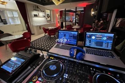 Poste de DJ avec platines, table de mixage et ordinateurs dans un salon élégant avec fauteuils rouges et éclairage tamisé.
