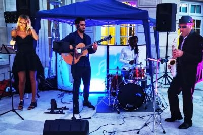 Un groupe de musiciens joue en plein air sous une tente, avec une chanteuse, un guitariste, un batteur et un saxophoniste.