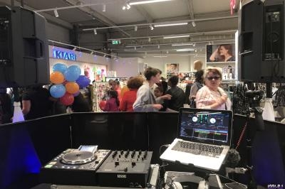 Poste de DJ avec platines et ordinateur dans un magasin, des clients et des ballons visibles en arrière-plan.