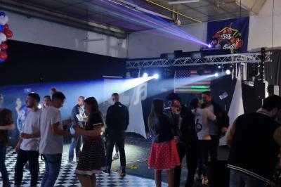 Des personnes dansent dans une salle décorée avec des ballons, des lumières colorées et un drapeau américain en arrière-plan.