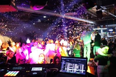 Personnes dansant sous des lumières colorées et des bulles dans une ambiance festive, vue depuis une table de DJ.