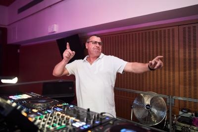 Un homme en polo blanc danse derrière une table de mixage dans une salle éclairée par des lumières colorées.