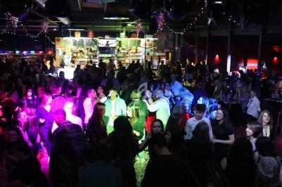 Salle de fête animée avec des personnes dansant sous des lumières colorées, un bar lumineux en arrière-plan.