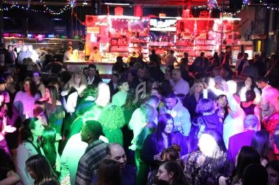 Foule dansante dans une salle de fête éclairée par des lumières colorées, avec un bar illuminé en arrière-plan.