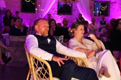 Un couple assis sur des chaises en rotin, tenant leurs mains, lors d'une réception éclairée par des lumières violettes.