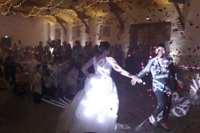 Un couple danse sous des lumières et des pétales rouges, entouré d'invités dans une salle décorée de guirlandes lumineuses.