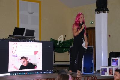 Une femme avec une perruque rose parle au micro sur une scène, près d'un écran affichant une photo d'anniversaire.