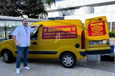 Un homme souriant se tient près d'un véhicule utilitaire jaune marqué ’Sound Light’, avec du matériel audio visible.