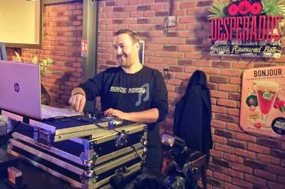 Un DJ souriant mixe de la musique sur un ordinateur portable dans un bar à la décoration murale en briques.