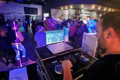 Un DJ mixe de la musique dans un bar animé avec des lumières colorées et des personnes dansant en arrière-plan.