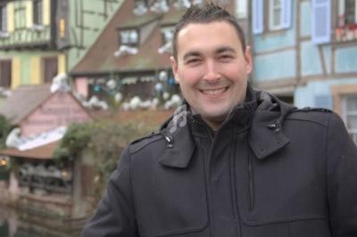 Homme souriant en manteau noir devant des maisons à colombages colorées dans un décor urbain hivernal.
