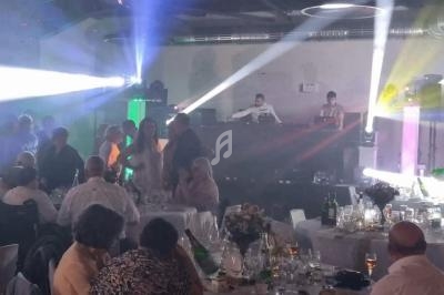 Des convives assis et debout dans une salle de réception éclairée par des jeux de lumière, avec un DJ en arrière-plan.