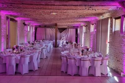 Salle de réception décorée pour un mariage, avec tables rondes dressées et éclairage rose sur les murs en pierre.