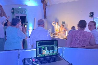Des invités applaudissent autour d'un couple devant un gâteau de mariage, avec une table de DJ au premier plan.
