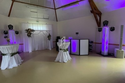 Salle de réception décorée avec des tables hautes drapées, un rideau blanc fleuri et un éclairage violet.