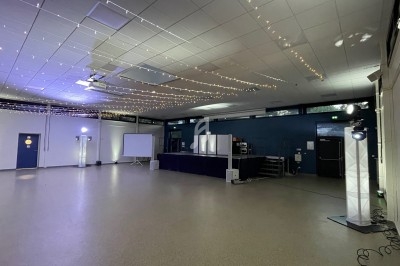 Salle vide avec scène, projecteurs, guirlandes lumineuses au plafond et écran de projection sur le côté.