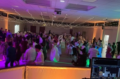 Salle de réception décorée avec des guirlandes lumineuses, des invités dansant et une console de DJ au premier plan.