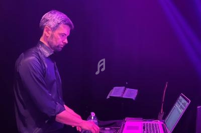 Un homme utilise une table de mixage et un ordinateur portable sur une scène éclairée par des lumières violettes.