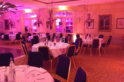 Salle de réception élégante avec tables rondes dressées, chaises bleues et éclairage rose tamisé.