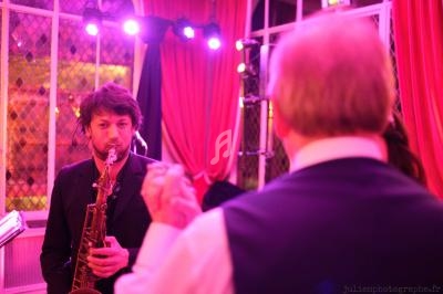 Un musicien joue du saxophone dans une salle éclairée de lumières roses, face à une personne de dos.