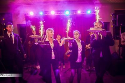 Serveurs en tenue formelle portant des plateaux avec des desserts et des cierges magiques dans une ambiance festive.