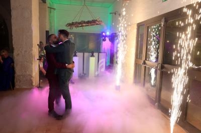 Deux hommes dansent enlacés sur une piste entourée de fumée, lumières colorées et étincelles en arrière-plan.