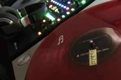 Platine vinyle avec disque rouge en gros plan, casque audio et table de mixage éclairée en arrière-plan.