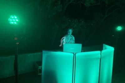 Un DJ mixe de la musique derrière une table lumineuse verte, entouré de projecteurs dans un environnement extérieur nocturne.