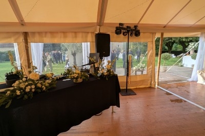 Intérieur d'une tente avec une table décorée de fleurs blanches, donnant sur un jardin où des personnes sont rassemblées.
