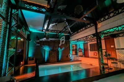 Salle industrielle rénovée avec une piscine intérieure, éclairage coloré et structures métalliques apparentes.