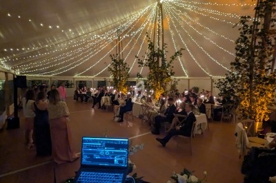 Salle de réception sous une tente éclairée par des guirlandes lumineuses, avec des invités assis et un poste de DJ au…