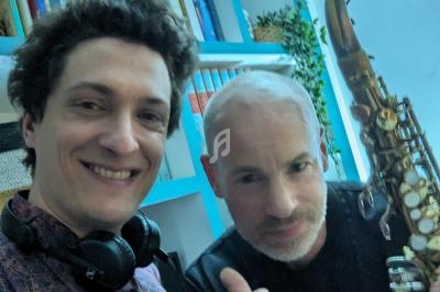 Deux hommes souriants posent dans une pièce avec des étagères, l'un tenant un saxophone.