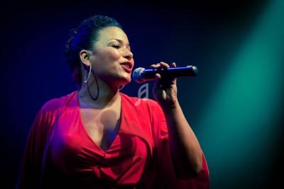 Femme en robe rouge chantant sur scène avec un micro, éclairée par une lumière bleue et verte.