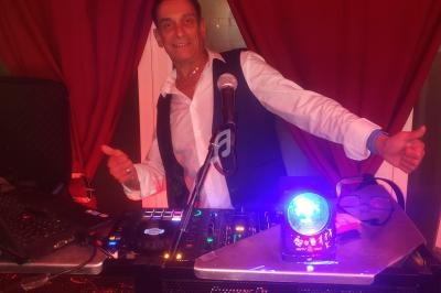 Homme souriant devant une table de mixage, entouré de rideaux rouges et éclairé par une lumière colorée.