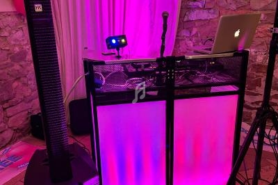 Poste de DJ avec éclairage coloré rose et violet, micros, ordinateur portable et enceintes dans une salle en pierre.