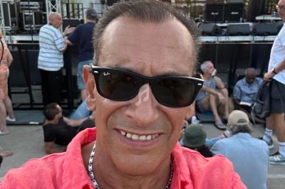 Homme en chemise rose et lunettes de soleil prenant un selfie devant une scène en plein air au coucher du soleil.