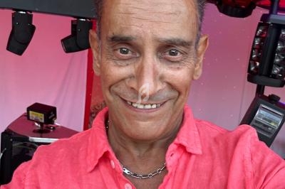 Homme souriant portant une chemise rose, se tenant devant un équipement de sonorisation sous un éclairage rouge.