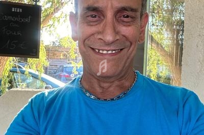 Homme souriant portant un t-shirt bleu, debout en extérieur près d'une fenêtre avec un tableau noir en arrière-plan.