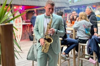 Un homme en costume vert joue du saxophone dans un bar en plein air, entouré de clients assis et debout.