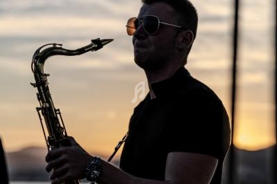 Un homme jouant du saxophone au coucher du soleil, portant des lunettes de soleil et une chemise noire.
