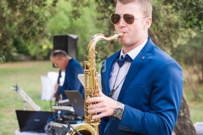 Un musicien en costume bleu joue du saxophone en extérieur, avec un autre musicien et du matériel audio en arrière-plan.