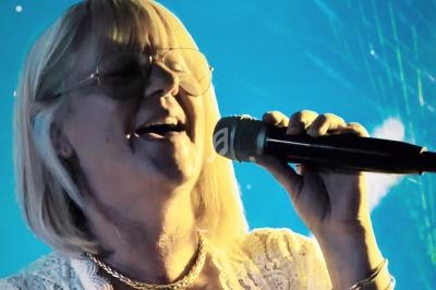 Femme blonde portant des lunettes, chantant au micro sur une scène éclairée de lumières bleues.