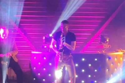 Un musicien joue du saxophone sur une scène éclairée par des lumières colorées, avec un panneau ’Venice Gym’ en arrière-plan.