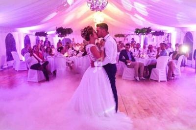Un couple danse leur première danse de mariage dans une salle décorée avec un éclairage violet et un sol recouvert de fumée.