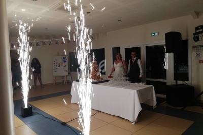 Un couple en tenue de mariage se tient près d'une pièce montée et d'une fontaine de verres, entouré de jets d'étincelles.